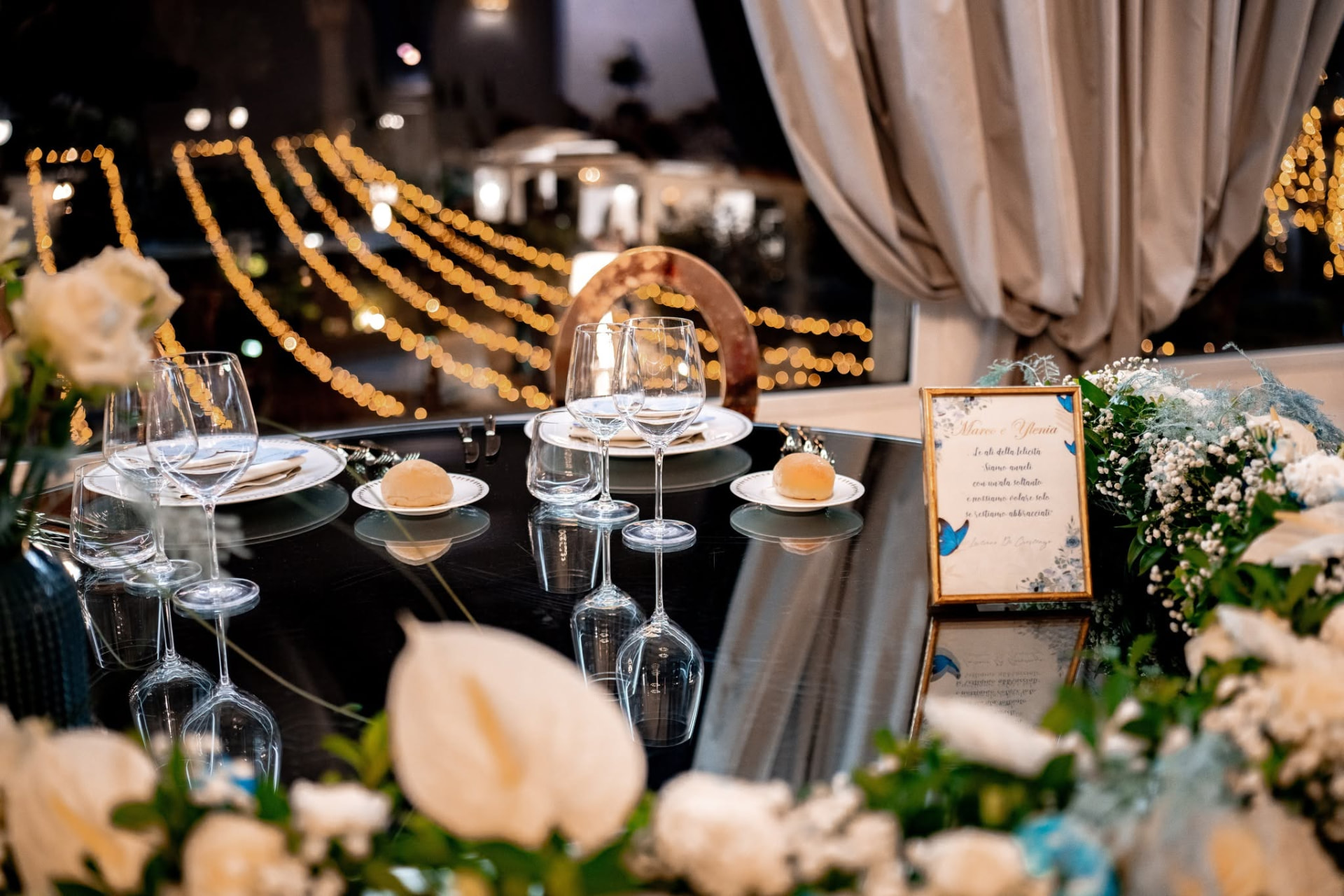 Il tavolo degli sposi apparecchiato davanti alla finestra, con fiori bianchi, il menu d'onore e le lucine fuori dal vetro