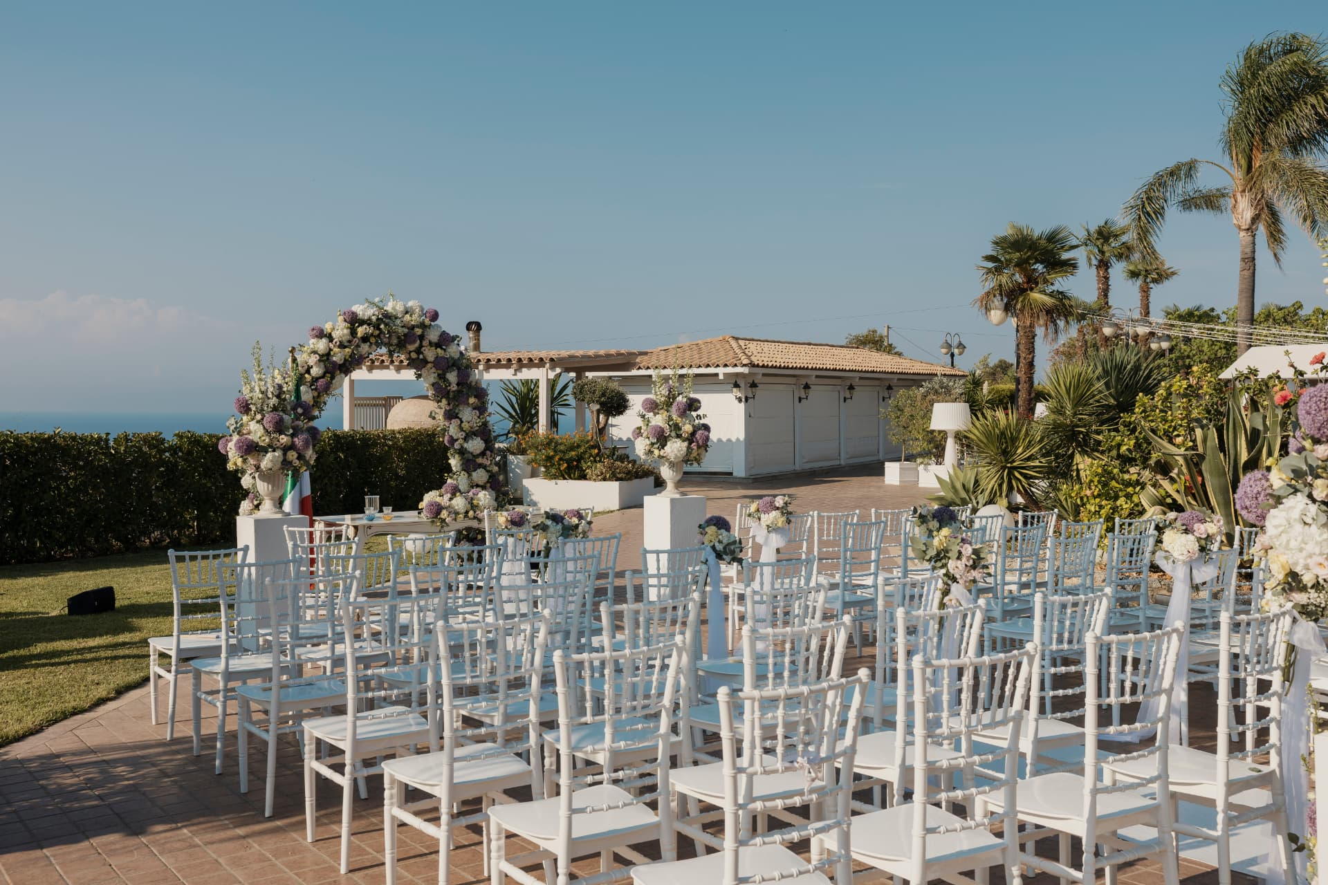 L'allestimento per il rito all'aperto di Villa Perara, con file di sedie bianche, arco floreale e il mare sullo sfondo